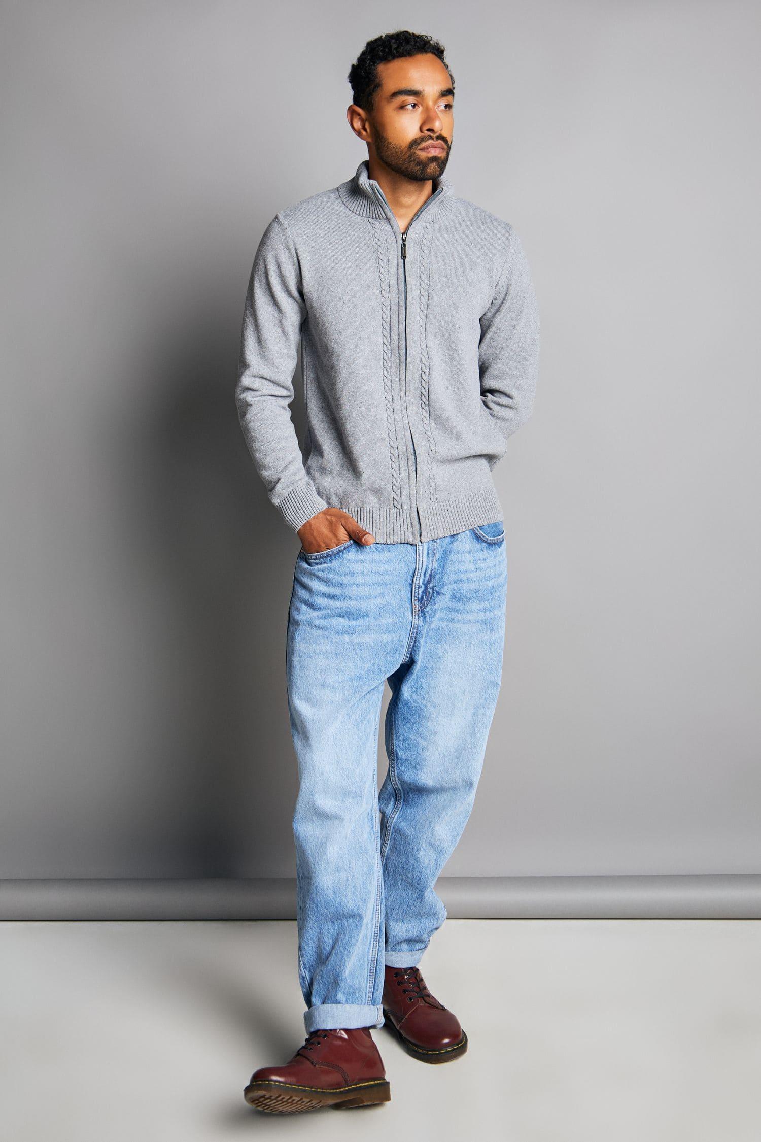 Hombre con suéter gris tejido de punto, jeans claros y botas marrón en fondo gris